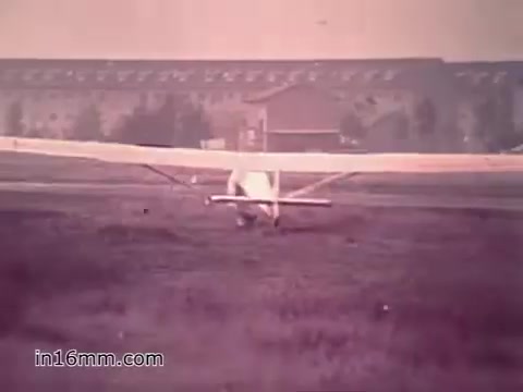 The still from the PSA 'Your Man in Nürnberg' features a white aircraft on an airfield, likely in a military setting. In the background, a large building with multiple windows is visible, indicating a base environment. The tone of the image has a vintage, slightly faded quality typical of 1960s film. This scene reflects the combination of military duties and leisure activities, highlighting the life of Specialist Four Joe Naifus as he balances his responsibilities in supply with opportunities for sightseeing.