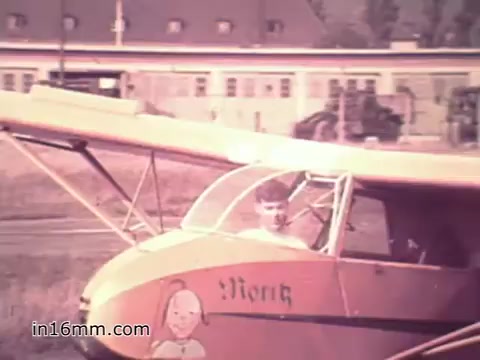 The still from the 1960s US Army recruiting PSA 'Your Man in Nürnberg' features a young soldier, likely Joe Naifus, inside a light aircraft. The plane is brightly colored and appears to be on a grassy field with military buildings in the background. The scene conveys a sense of adventure and highlights the soldier's role in the Army, focusing on both his professional responsibilities and leisure activities like flying.