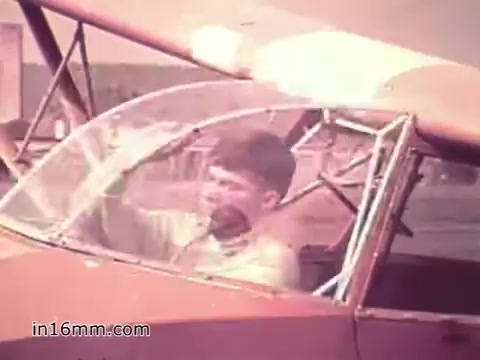The still from the 1960s PSA 'Your Man in Nürnberg' portrays a young soldier, Joe Naifus, in the cockpit of a small red aircraft. He appears focused, likely preparing for takeoff or checking instruments. The background suggests an airfield setting, emphasizing Joe's role within the U.S. Army related to supply and logistics. The imagery reflects the blend of military duty and personal exploration, highlighting the multifaceted life of a soldier stationed in Nuremberg.