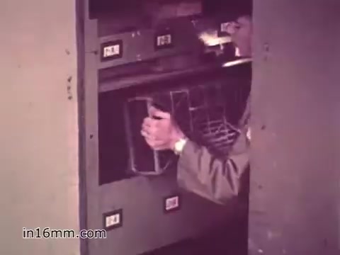 The still from the PSA 'Your Man in Nürnberg' shows a soldier, identified as Joe Naifus, engaged in his supply duties within a military setting. He is seen retrieving items from a storage unit, highlighting the meticulous nature of his job in managing various parts necessary for operations. The scene conveys a sense of responsibility and efficiency, emphasizing Joe's skills learned through Army training. The backdrop hints at a bustling environment, reflecting the hectic yet structured life of a U.S. Army specialist in the 1960s.