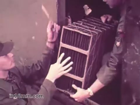 The still from the 1960s US Army recruiting PSA 'Your Man in Nürnberg' depicts two soldiers engaged in handling a large, rectangular object, likely a piece of equipment or supply part. One soldier, seated, uses both hands to stabilize the object, while the other, standing, supports it from below. Their uniforms indicate military service, and the setting appears to be a maintenance or supply environment, emphasizing the logistical aspect of Army operations. The color tone suggests a vintage look typical of that era.