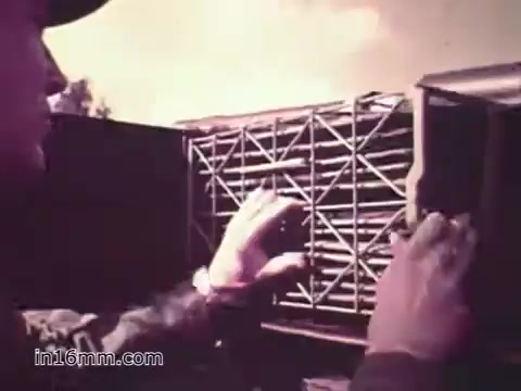 The still from the PSA 'Your Man in Nürnberg' features a Specialist Four, Joe Naifus, engaged in his supply job with the United States Army. He is shown handling a large stack of supplies or equipment parts, emphasizing the complexity and responsibility of his role. The image captures a moment of focus and diligence, portraying the hectic but rewarding nature of military logistics. The background suggests an outdoor setting, hinting at Joe's work-life balance, which includes sightseeing in Nuremberg.