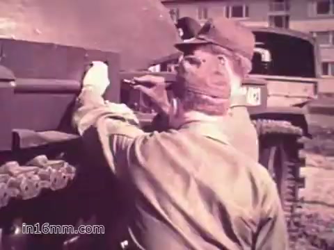 The still from 'Your Man in Nürnberg' depicts two U.S. Army soldiers working on a military vehicle. One soldier, identified as Joe Naifus, is focused on a specific part of the vehicle, while the other assists him. Both are in uniform, indicating their military roles. The background features a military installation, suggesting the setting is a base in Nuremberg. The scene emphasizes the technical and collaborative nature of their work, highlighting the skills learned in the Army. The tone conveys a sense of duty and professionalism.