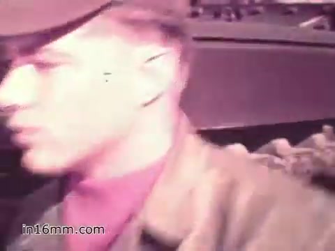 The still from the 1960s PSA 'Your Man in Nürnberg' features a U.S. Army soldier, identified as Specialist Four Joe Naifus, in a military uniform. He appears focused and determined, likely on duty. In the background, elements of military equipment can be seen, hinting at his role in supply management. The scene conveys a sense of purpose, showcasing the hectic yet skilled nature of his job.