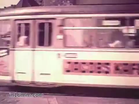 The still from 'Your Man in Nürnberg' features a 16mm shot of a public transportation tram, likely in Nuremberg, Germany. The tram is painted in a light color with advertisements visible on its side. The background suggests a cityscape typical of the 1960s, with buildings and a hint of street life. This scene emphasizes the everyday life aspect experienced by soldiers like Joe Naifus, highlighting both their military responsibilities and their leisure time exploring the local culture.