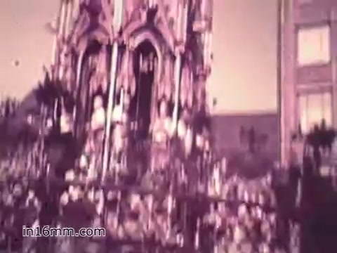 The still from the PSA 'Your Man in Nürnberg' features a busy scene, likely showcasing a vibrant public area with many people gathered around an ornate building or monument. The faded colors suggest it’s a vintage 16mm film from the 1960s, capturing both the bustling atmosphere and the cultural backdrop of Nürnberg. The context emphasizes Joe Naifus's role in the U.S. Army, highlighting his competence in supply management while hinting at his off-duty experiences exploring the city.