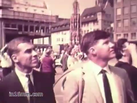 The 16mm still from the PSA 'Your Man in Nürnberg' features two men, likely soldiers, looking up and observing their surroundings, possibly in a city square. They are dressed in business-like attire, suggesting a mix of military professionalism and civilian exploration. The environment appears bustling, with a mix of people and buildings, indicating a vibrant setting in Nuremberg. This scene captures the dual life of military personnel: the responsibilities of their duties and the opportunity for leisure and exploration.