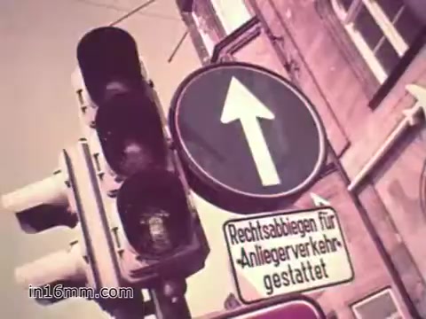 The still from the PSA 'Your Man in Nürnberg' features a traffic signal showing a red light alongside a circular sign with an upward arrow, indicating straight traffic. Below the sign, there's a message in German, likely related to traffic rules. The colors are slightly muted, characteristic of 16mm film. The image captures the urban environment of 1960s Nürnberg, reflecting the post-war setting and the daily life of U.S. Army personnel stationed there.