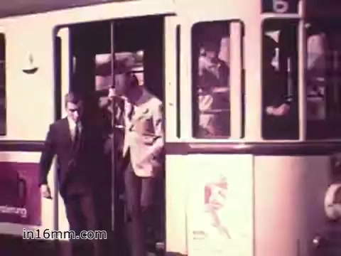 The still shows a scene from the 1960s PSA 'Your Man in Nürnberg,' featuring two men exiting a public transportation vehicle in Nuremberg. One man is dressed in a suit, while the other wears a lighter-colored jacket. The setting reflects a busy urban environment, indicative of military life balanced with leisure activities, as Joe Naifus, a Specialist in the U.S. Army, manages complex supply tasks during the day and enjoys sightseeing afterward.