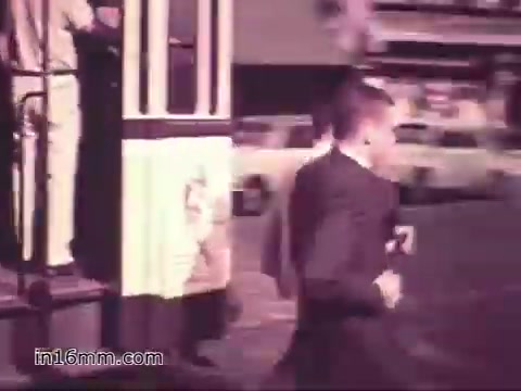 The still from 'Your Man in Nürnberg' features a young man in a suit, likely Joe Naifus, hurrying away from a tram or trolley stop. The setting appears lively, indicative of a bustling city environment, with cars visible in the background. This scene captures the essence of his day-to-day life as a soldier in the US Army, emphasizing both his professional responsibilities and the vibrant culture of Nuremberg that he explores after work.