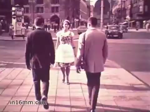 The still from the 1960s US Army recruiting PSA 'Your Man in Nürnberg' features two men in suits walking towards a woman in a white dress, who stands on the sidewalk. The setting appears to be a city street in Nuremberg, with buildings and vehicles in the background. The scene captures a moment of casual interaction, showcasing the vibrancy of daily life and the cultural context of the time.