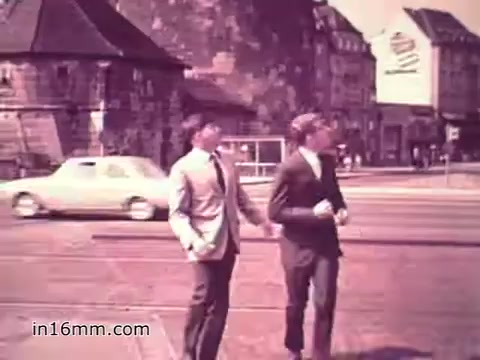The still from the 1960s US Army recruiting PSA 'Your Man in Nürnberg' features two young men in suits walking together on a city street. The backdrop includes a mix of traditional and modern buildings, with a car passing by and a large advertisement visible. The scene conveys a sense of camaraderie and exploration, reflecting the message of military service and personal growth.