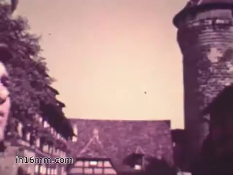 The still from the 1960s PSA 'Your Man in Nürnberg' features a US Army Specialist Four, Joe Naifus, likely depicted against a backdrop of historic architecture in Nuremberg. The composition suggests a blend of military life and personal exploration, showcasing the contrast between his structured job in supply and leisure activities like sightseeing. The overall tone seems nostalgic, capturing a moment of duty intertwined with personal experience in a significant locale.