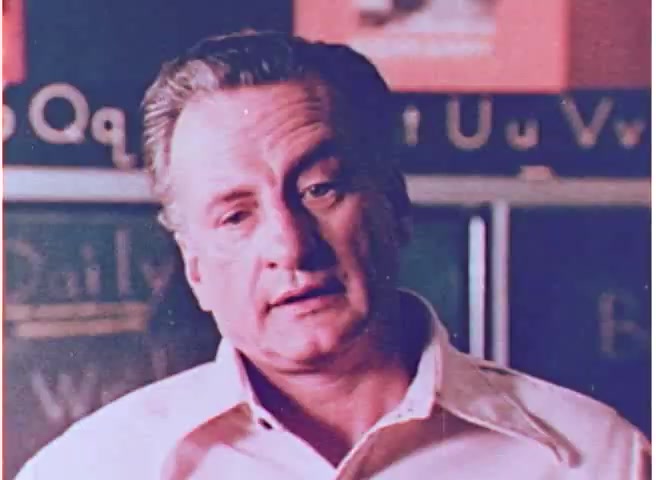 The still features George C. Scott in a thoughtful expression, likely addressing an audience. He wears a light-colored shirt and appears to be in a classroom setting, with chalkboards and educational materials in the background. The image evokes a sense of seriousness and urgency, aligning with the PSA's message about fuel conservation in the 1970s. The overall tone suggests a blend of authority and concern for the subject matter.