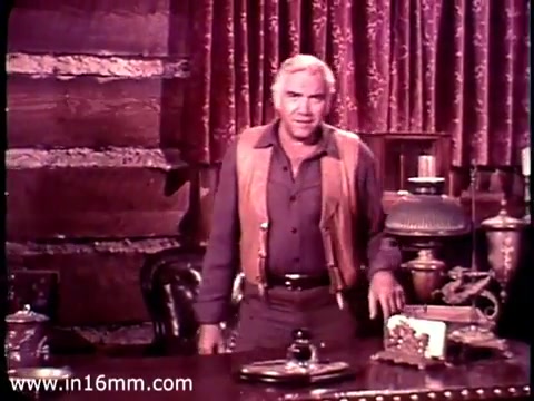 The still features Lorne Greene in a rustic, wooden-set environment, typical of his role in 'Bonanza.' He is dressed in a buttoned-down shirt with a vest, appearing engaged and earnest. The backdrop includes rich curtains and various props, suggesting a thoughtful atmosphere. Greene is likely addressing the audience, possibly reading from cue cards, as indicated by your brother's observation. The overall tone reflects a blend of authority and compassion, fitting for a public service announcement about overseas aid.