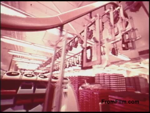The still from the 'Bell System Film from the 1970s' depicts a factory or assembly line setting. It features overhead conveyor systems with hanging apparatuses, likely used for transporting materials or products. The background shows stacks of equipment or containers, creating a colorful and busy atmosphere typical of industrial environments from that era. The lighting gives the scene a warm tone, accentuating the machinery and operational aspects of the facility.