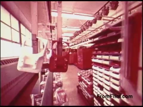 The still from the 'Bell System Film from the 1970s' depicts an industrial setting, featuring an organized storage area with a variety of tools and equipment. In the foreground, there are white and red tool carriers or carts hanging from a rail. The background showcases shelves filled with boxes, suggesting a well-maintained workshop atmosphere typical of the telecommunications industry during that era. The overall color palette is slightly muted, characteristic of older film stock.