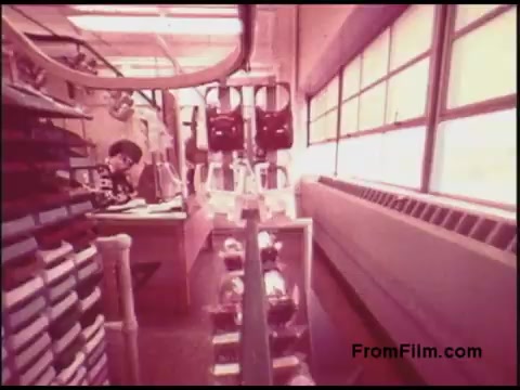 The still from the 1970s Bell System film depicts an interior of an office or lab environment, characterized by large windows allowing natural light to flood in. In the foreground, there are shelves filled with boxes or equipment, while a person is seated at a desk, seemingly engaged in work or research. The overall atmosphere suggests a mix of technology and traditional office elements, typical of the era. The light has a warm hue, contributing to the nostalgic look of the film.
