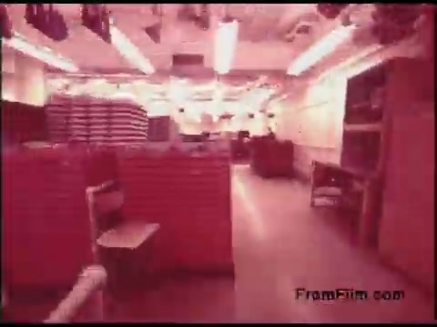 The still from the 1970s Bell System Film depicts an indoor space, likely a storage or equipment room, with stacked materials or equipment in the background. The lighting is bright, suggesting a utilitarian environment. Red or pink hues dominate the image, indicating possible color degradation typical of older film stock. The layout features sparse furniture, including a chair and a table, contributing to a functional, industrial aesthetic.