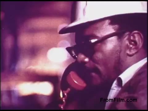 The still depicts a man wearing glasses and a hard hat, speaking into a red handheld telephone. He appears to be in an industrial or construction environment, with blurred lights in the background suggesting action and activity. The color tones are warm, characteristic of 1970s film aesthetics. The image captures a moment of communication in a professional setting, reflecting the era's focus on technology and infrastructure.