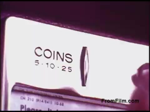 The still features a close-up view of a coin slot or mechanism, indicating denominations of 5, 10, and 25 cents. It's likely part of a vending or payphone system, commonly used in the 1970s. The film captures a nostalgic moment, emphasizing the era's technology and consumer interaction with coin-operated machines. The aesthetic reflects the film's vintage style, characteristic of that period.