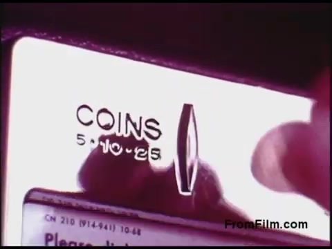 The still features a close-up view of a vintage coin slot, likely from a payphone or similar device, with the word 'COINS' prominently displayed. Below it, denominations of 5, 10, and 25 cents are marked. A hand is shown about to insert a coin, conveying a sense of interaction with the machine. The colors appear to have a retro quality, characteristic of 1970s film, enhancing the nostalgic feel of the scene.