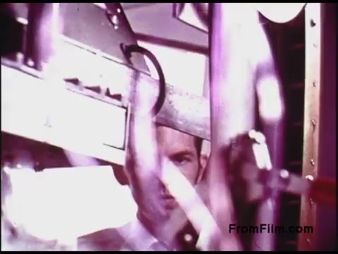 The still from the 'Bell System Film' shows a close-up of machinery, likely related to telecommunications or film processing from the 1970s. A man's face is partially visible, hinting at a connection to the technology being operated. The colors are slightly faded, typical of older film stock, and the focus on the machinery suggests an emphasis on technical processes or innovation during that era.