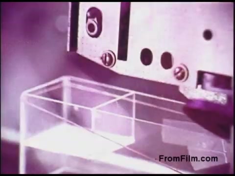 The still from a 16mm Bell System film from the 1970s depicts a close-up of a mechanical device possibly engaged in a technical process. The focus is on the device's components, including screws and an aperture, positioned above a transparent container. The overall color tone leans towards a purple hue, suggesting a vintage aesthetic typical of that era’s film quality. The image captures a moment of industrial or laboratory activity, reflecting the technological advancements of the time.