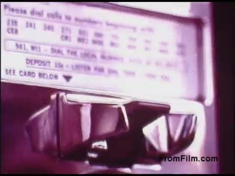 The still features a close-up of a vintage payphone from the 1970s, showcasing its metal details and keypad. The payphone displays instructions for dialing numbers, including area codes, and emphasizes deposit requirements for calls. The colors and design reflect the aesthetic common in public telecommunication devices of that era, highlighting the transitional phase of technology.