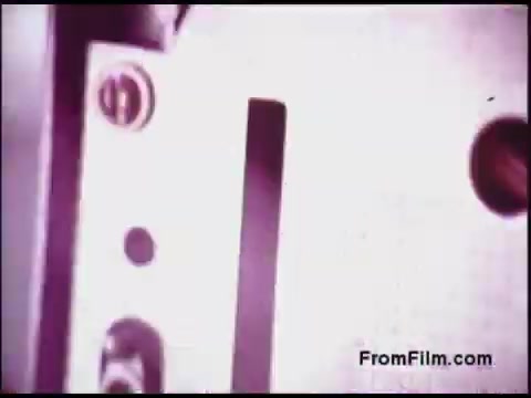 The still appears to show a close-up of a mechanical device, likely related to film processing or telecommunications, typical of the 1970s technology context. The image features various metallic components, including a slot or opening, and possibly some controls or indicators, reflecting the industrial design of that era. The color palette suggests a faded or vintage look, which adds to its historical significance.