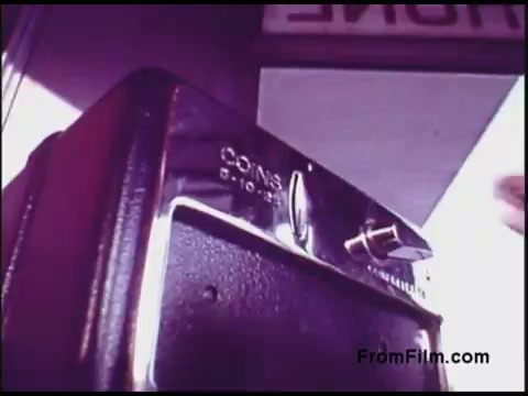 The still depicts a close-up of a vintage payphone from the 1970s, prominently featuring a coin slot labeled 'COINS' with denominations of 5, 10, and 25 cents. The angle suggests a focus on the act of inserting coins, emphasizing the retro technology used for telephone calls during that era. The background appears blurred with hints of signage, contributing to the nostalgic atmosphere.
