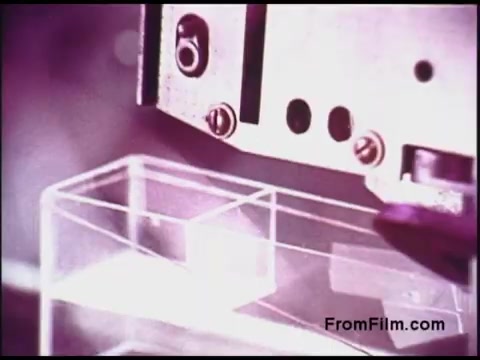 The still from the 16mm 'Bell System Film from the 1970s' depicts a close-up view of a mechanical apparatus, likely part of a telecommunications or technology demonstration. The device appears to be interacting with a transparent, cubic container, showcasing intricate details of machinery and emphasizing precision. The film's color palette suggests a vintage look, typical of the era, with a focus on technical processes.