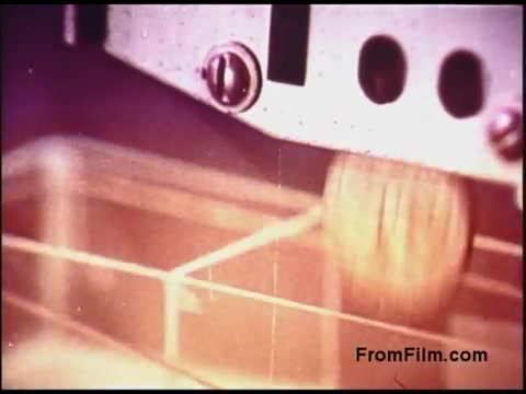 The 16mm still from the 'Bell System Film from the 1970s' showcases a close-up view of a machinery component. In the image, a round object appears to be falling or rolling towards a structured area, possibly within a production or sorting mechanism. The color palette reflects the vintage quality of the film, likely featuring muted tones typical of that era. The overall aesthetic evokes a sense of industrial design and the technological advancements of the time.