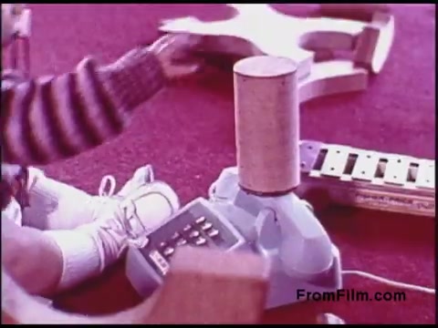 The 16mm still from the 'Bell System Film from the 1970s' features a child's hand reaching towards a toy device, likely a learning phone or similar gadget. The background is carpeted, with colorful toys scattered around, including a xylophone and other wooden shapes, reflecting a playful and educational environment. The child appears to be engaged in play, showcasing early childhood learning through interaction with toys.