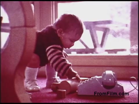 The still shows a young child, possibly a toddler, kneeling on a vibrant carpet while reaching towards a retro-style telephone. The child is dressed in a striped sweater and white socks, exuding a playful curiosity. The setting appears to be a cozy interior with natural light filtering in through a nearby window, highlighting the nostalgic atmosphere typical of the 1970s. The scene captures a moment of innocence and exploration, characteristic of early childhood.