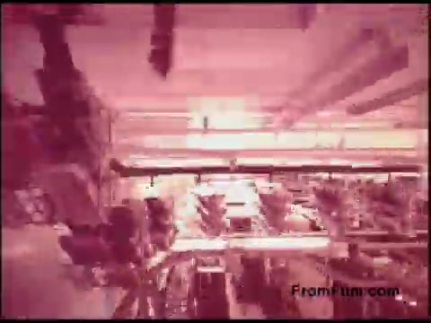 The 16mm still from the 'Bell System Film from the 1970s' captures an industrial setting, likely within a telecommunications or manufacturing facility. The image features an array of machinery and structures, presented with a reddish tint that may suggest a vintage aesthetic. The composition hints at a complex network of equipment, possibly showcasing the inner workings of telecommunication systems or production processes during that era. Overall, it reflects technological advancement and the operational environment of the time.