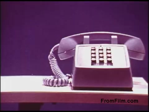 The still depicts a vintage telephone from the 1970s, set against a plain background. The phone, featuring a classic rotary design and a curly cord, sits on a simple wooden table. Its visible buttons and overall design reflect the technology of that era, emphasizing the focus on communication devices in the film. The image conveys a sense of nostalgia, highlighting a time when such telephones were commonplace.