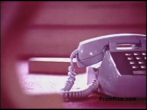 The still features a light blue rotary telephone sitting on a surface, likely a desk, with visible telephone cords. The color palette is soft and muted, characteristic of film from the 1970s, and the background appears slightly out of focus, emphasizing the phone as the focal point. This image likely illustrates communication technology of the era, possibly within a context related to the Bell System.
