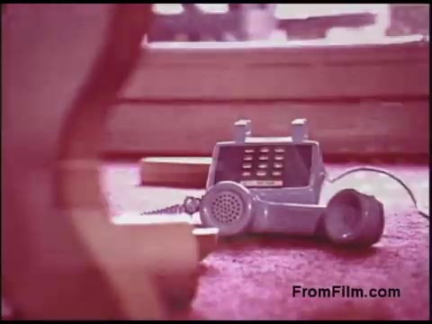 The still features a vintage gray rotary-style telephone sitting on a carpeted surface, indicative of the 1970s. The image has a soft focus and warm color tones, conveying a nostalgic ambiance. The phone is partially obscured, suggesting a casual, domestic setting, typical of the era's communication technology.