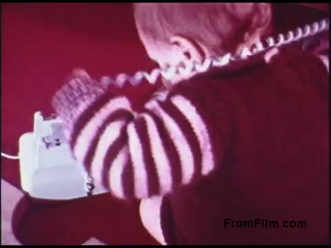 The still depicts a child, likely a toddler, sitting on a carpet, engaged with a classic rotary telephone. The child is wearing a striped sweater, which adds a vibrant touch to the scene. The film has a warm, nostalgic color palette typical of the 1970s, reflecting a playful yet simple interaction with technology from that era. The child's concentration and curiosity suggest an early encounter with communication devices.