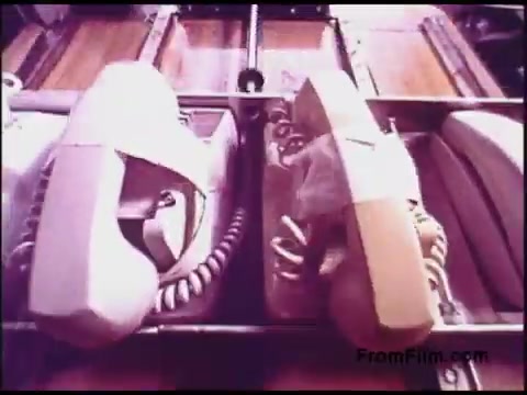 The still from the 'Bell System Film from the 1970s' features a close-up view of several retro telephones, arranged side by side. The phones are in soft colors, likely pastel shades, and are positioned on a production line, suggesting a manufacturing or assembly process. The background includes a hint of machinery, emphasizing the industrial setting. This imagery captures the era’s focus on telecommunication technology and its evolution.