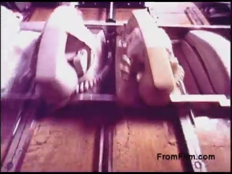 The still from the 'Bell System Film from the 1970s' features a close-up view of vintage telephones positioned on a track or conveyor system. The phones appear to be transitioning or being processed, showcasing an era of technology focused on telecommunications. The color palette reflects the muted tones typical of films from that period, adding a nostalgic feel to the imagery.
