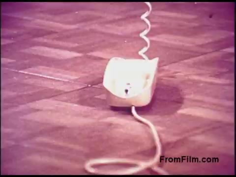 The still shows a close-up of a white telephone receiver resting on a wooden floor, with a coiled phone cord extending from it. The aesthetic has a vintage look, typical of the 1970s, featuring muted colors and a slightly faded appearance. The setting suggests a nostalgic context related to telecommunications from that era.