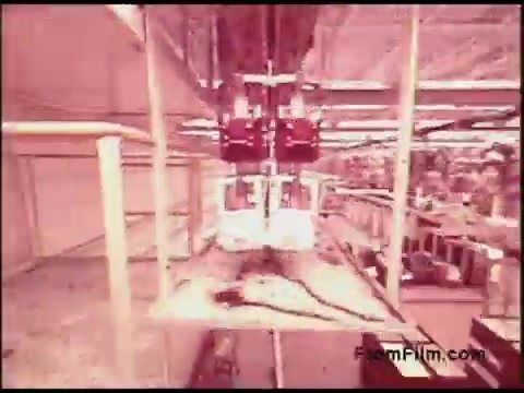 The still from the 'Bell System Film from the 1970s' presents an industrial scene, likely within a manufacturing facility. It features equipment suspended from above, possibly related to telecommunications or electronic assembly. The environment appears busy, with various components and machinery visible in the background, hinting at a focus on production processes typical for that era. The color tone suggests a vintage look characteristic of 16mm film.