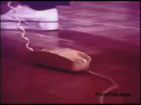 The still depicts a close-up of a vintage telephone on a wooden floor, with a coiled phone cord extending from it. The image is characterized by a warm color tone typical of 1970s film. A person's foot, wearing white shoes, is visible near the phone, suggesting an interaction with the device. The composition reflects a casual domestic scene likely related to communication.