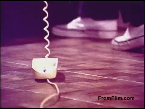The still features a close-up of a vintage telephone on a wooden floor, with a curly cord leading away from it. The telephone is yellow and has a playful, rounded design, typical of the 1970s aesthetic. In the background, a person's feet in white sneakers are partially visible, suggesting a casual, domestic setting. The overall color tone is warm, giving a nostalgic feel.