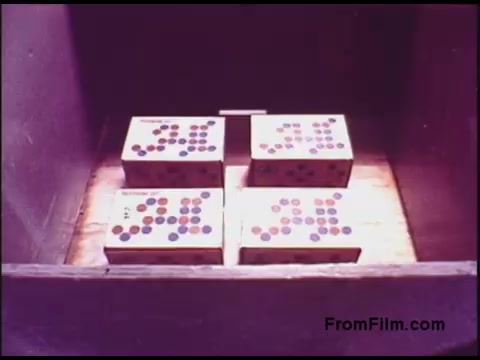 The still features a wooden box containing four rectangular packages. Each package is labeled with various colored dots arranged in patterns. The scene reflects a mid-20th-century aesthetic, typical of industrial or educational films from the 1970s, likely highlighting telecommunications technology or products related to the Bell System. The lighting is soft, emphasizing the textures of the box and the packages inside.