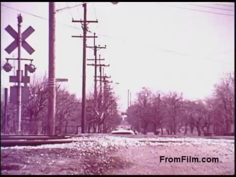 The still features a rural setting with a railroad crossing visible in the foreground. There are wooden utility poles along the side of the road, and the landscape is lined with trees, which appear bare, likely indicating early spring or late fall. The color palette has a faded quality typical of 16mm film from the 1970s, with muted tones that reflect the era's visual style. A vehicle is seen in the distance, suggesting a quiet, everyday scene.