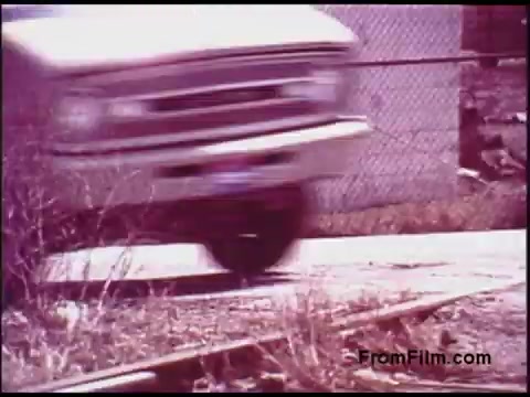 The still captures a motion blur of a vintage truck driving along a gravel road, with a chain-link fence in the background. Surrounding vegetation suggests a somewhat neglected environment. The pinkish hue indicates the color grading typical of 1970s film stock, evoking a nostalgic atmosphere. The focus is on the truck's movement, indicating a dynamic scene in the context of the film.