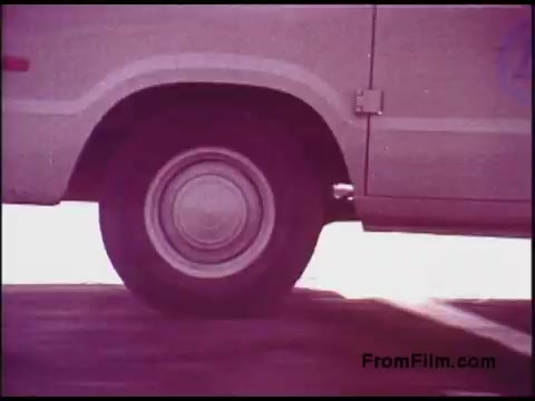 The still from the 'Bell System Film from the 1970s' features a close-up of a van's wheel on a road. The focus highlights the tire in motion, suggesting movement and activity. The color palette exhibits a vintage tone, typical of films from that era, evoking a sense of nostalgia. It captures a brief moment that reflects the transportation aspect of the Bell System's operations.